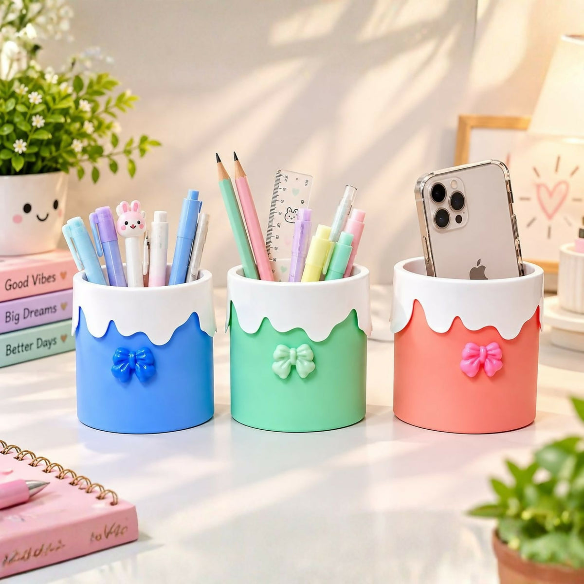 Three pastel organizers on a desk, filled with writing instruments and a phone, beside a pink notebook.