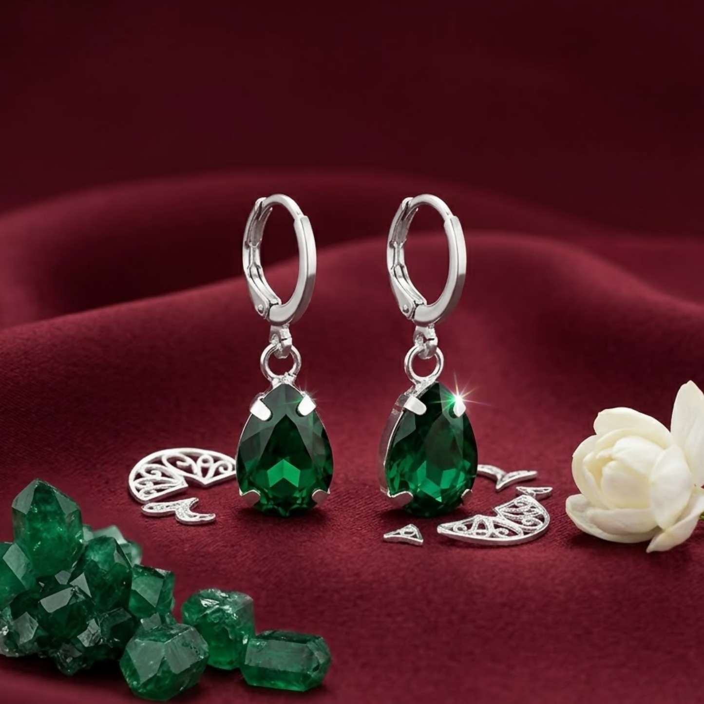 Green teardrop crystal earrings with silver hooks on a red satin background, with loose green crystals and a white flower.