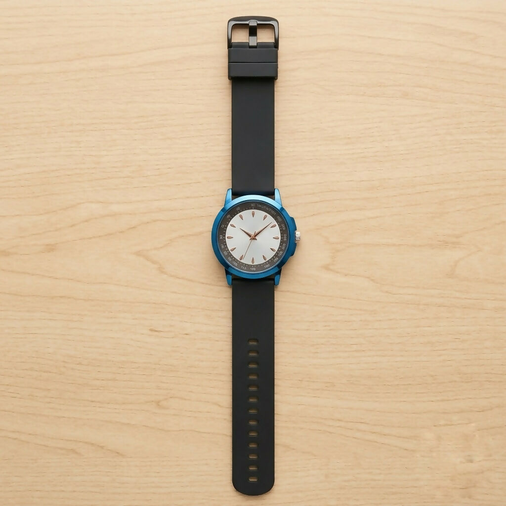 A blue watch with a white face, gold hands, and a black strap is viewed from above on a light wood surface.