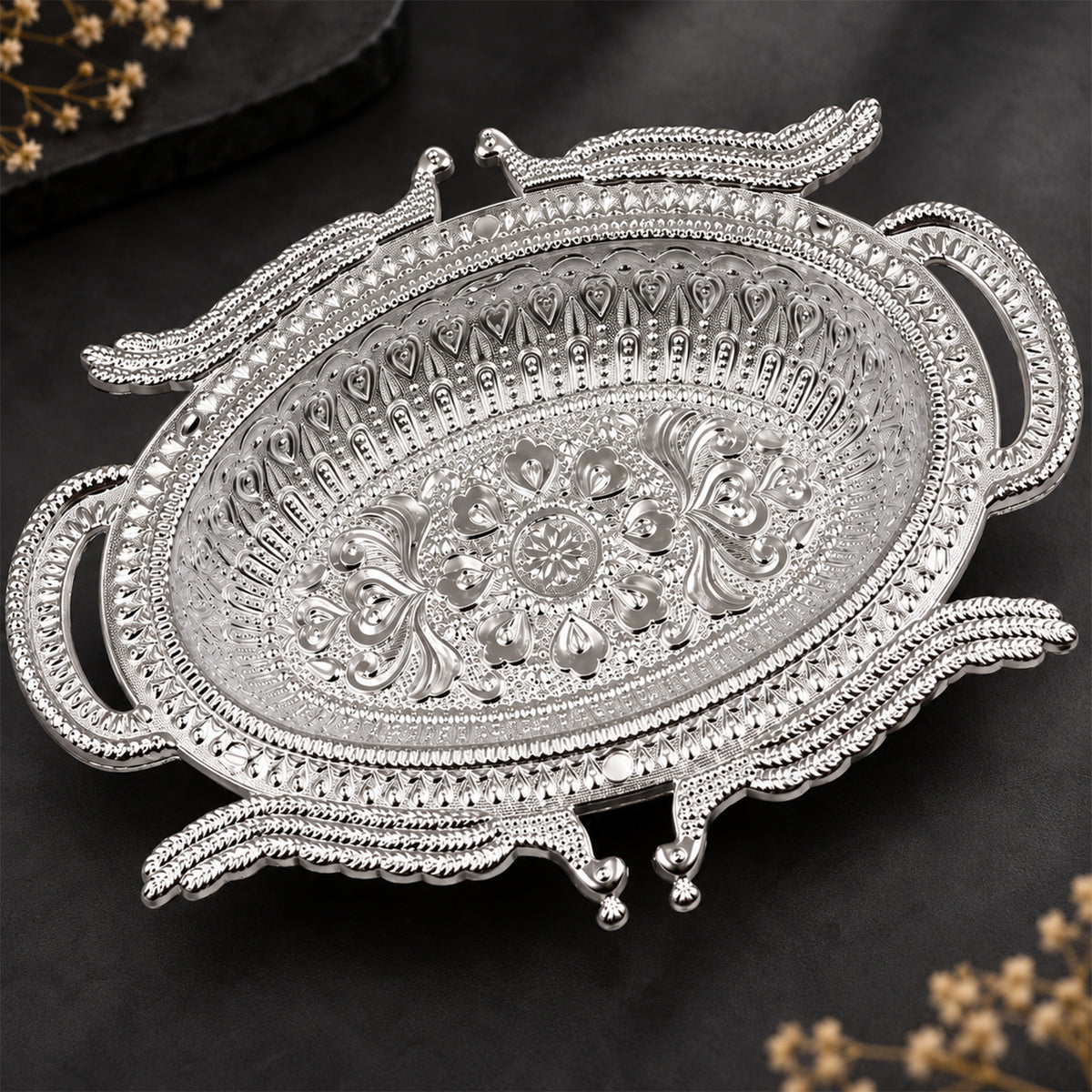 Empty silver-colored ornate serving tray with detailed patterns on a dark background with blurred floral elements.