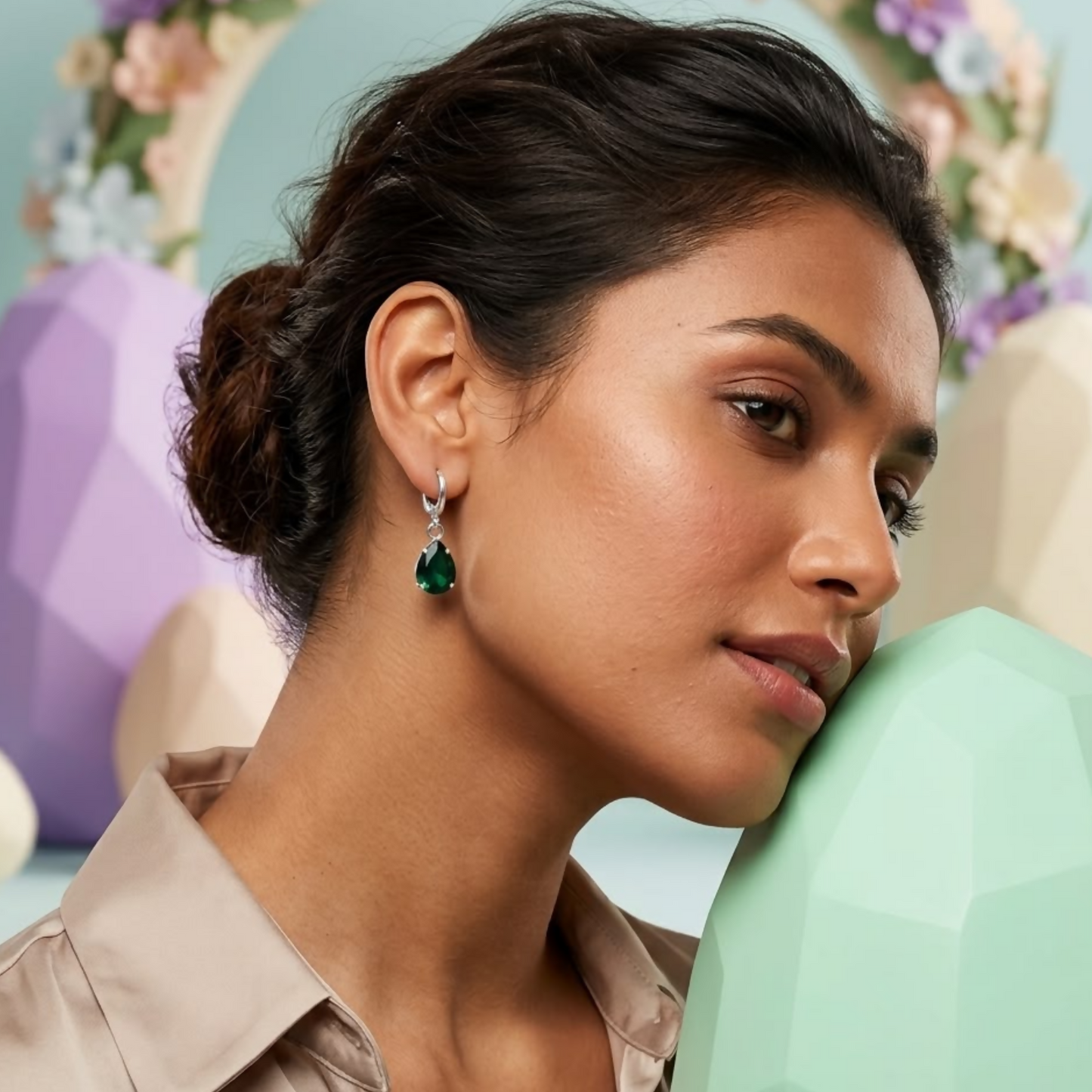 A woman with dark hair wears a single green teardrop crystal earring, looking thoughtful against a pastel background.