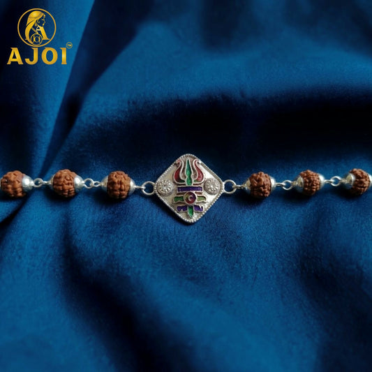 A silver-plated rakhi bracelet with brown rudraksha beads and a colorful trishul charm on a blue fabric background.