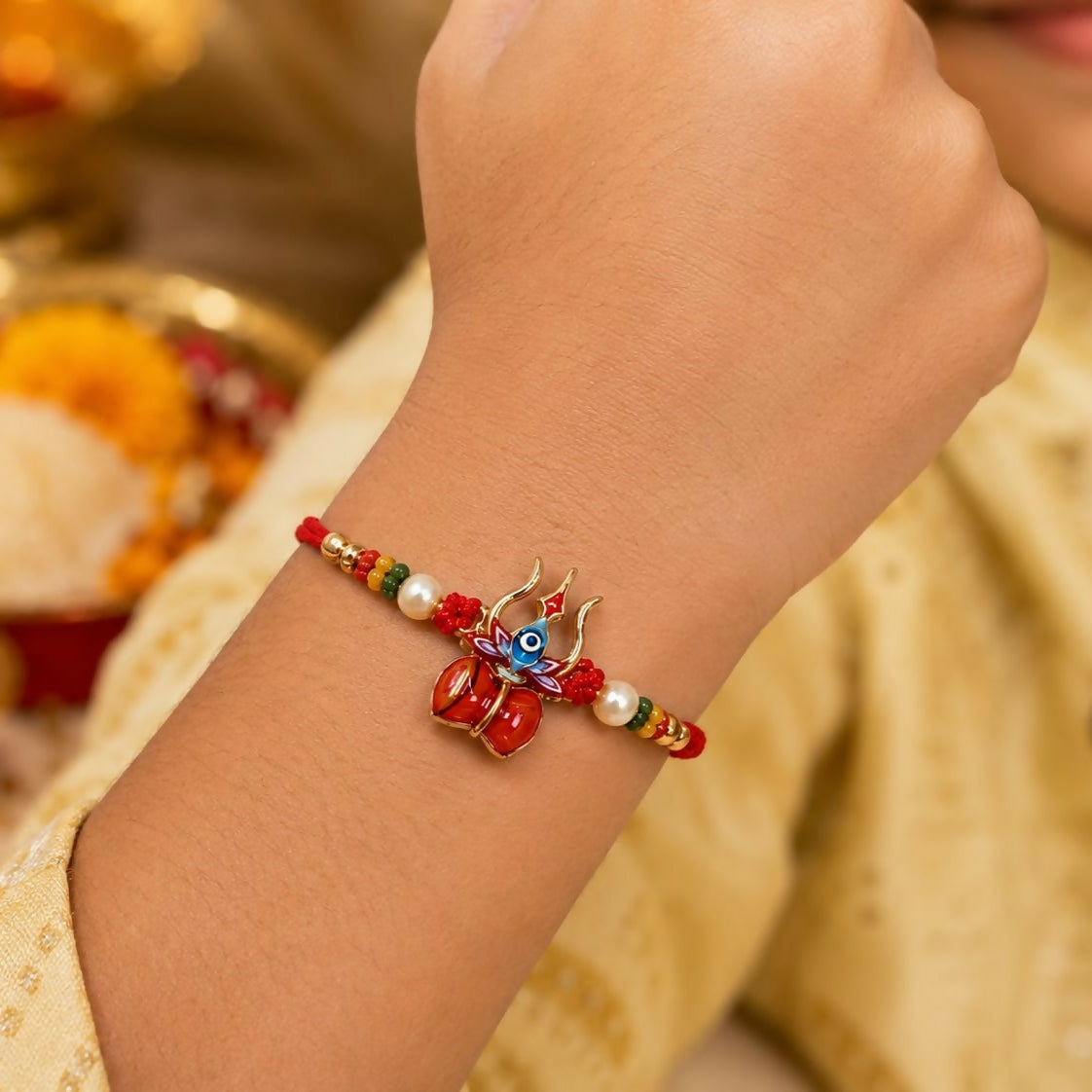A child's arm wears a red thread bracelet featuring a gold trishul and damru charm with blue enamel, flanked by pearls and