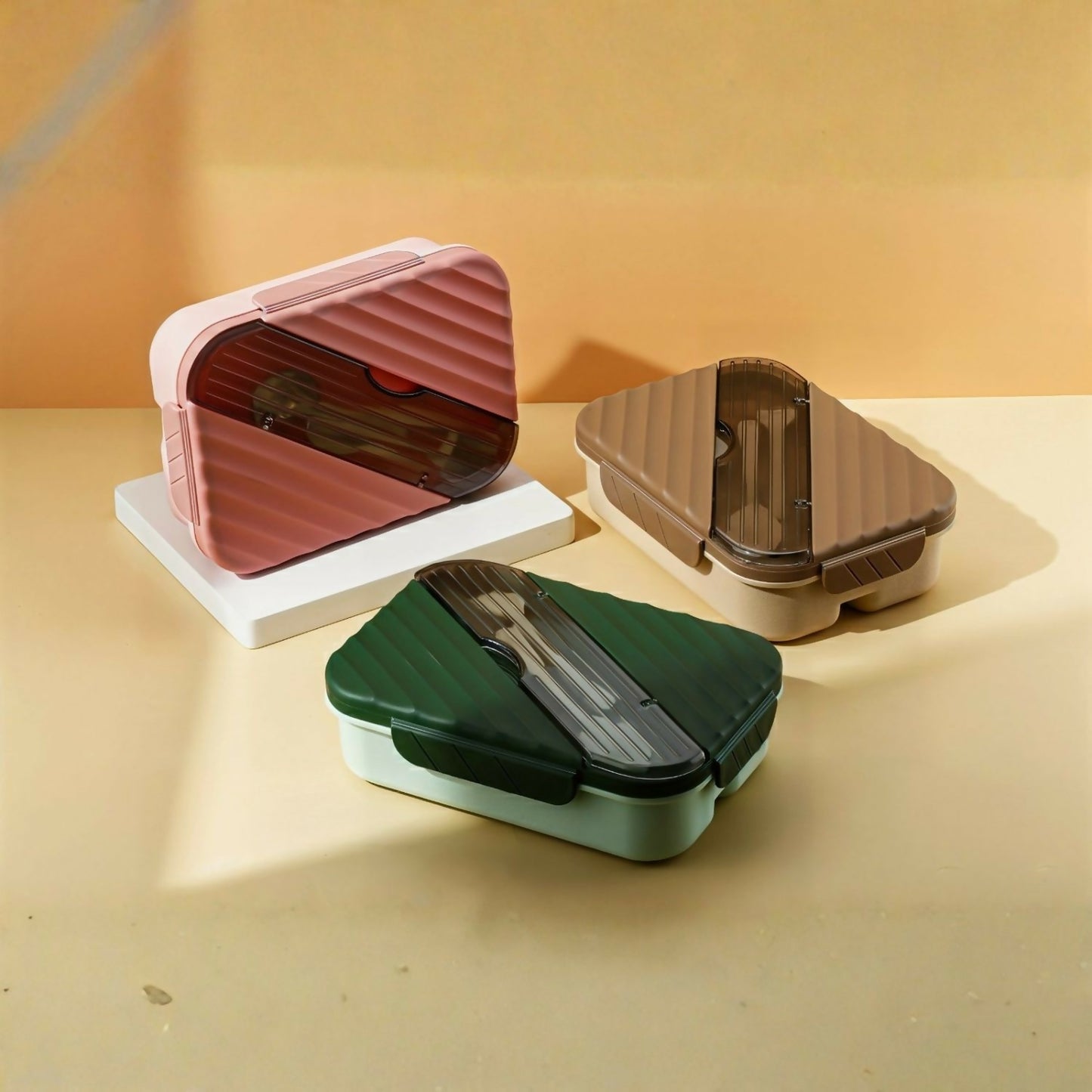 Three closed bento lunch boxes in pink, brown, and green with clear lids revealing cutlery, sitting on white and beige