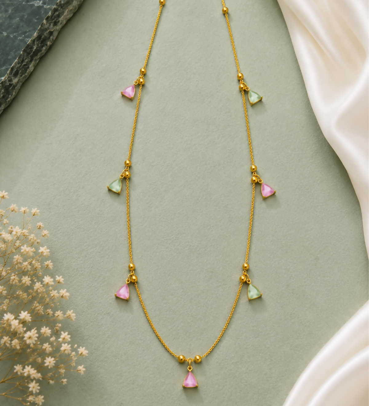 A gold-plated necklace with pink and mint green triangular charms is displayed on a pastel green surface.