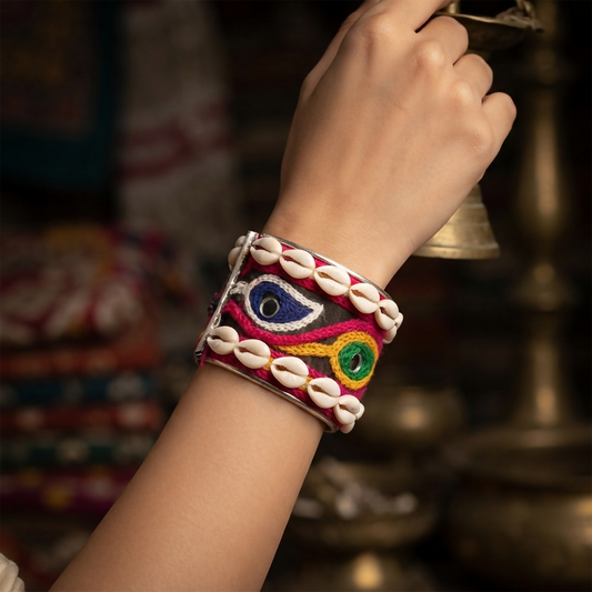 A person's arm wears a wide, silver-toned cuff bracelet adorned with colorful embroidery, mirrors, and white cowrie shells.