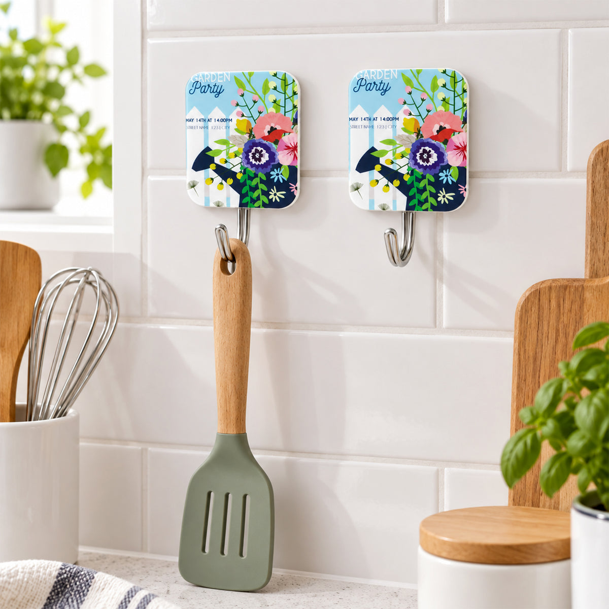Two floral-patterned adhesive hooks mounted on white kitchen tiles, one holding a green silicone spatula.