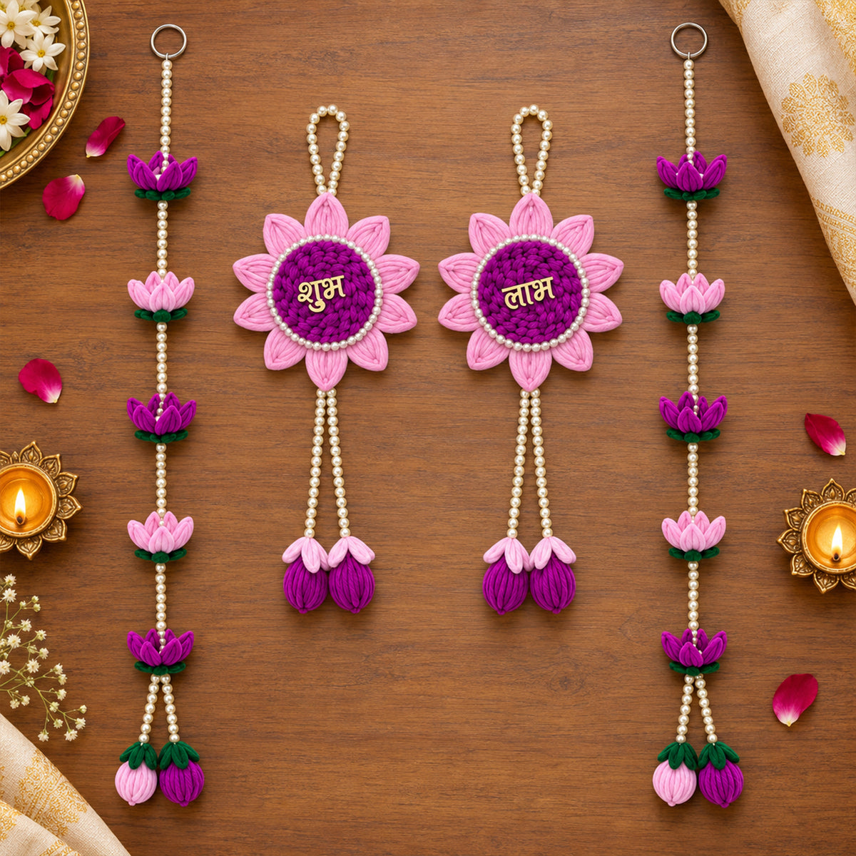 Two pink and purple lotus hangings with pearl beads and two lotus bead strings on a wooden surface with lit diyas.
