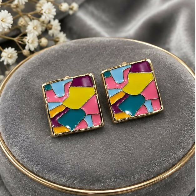 A pair of square stud earrings with abstract multicolour enamel design and gold-tone borders rest on grey velvet.
