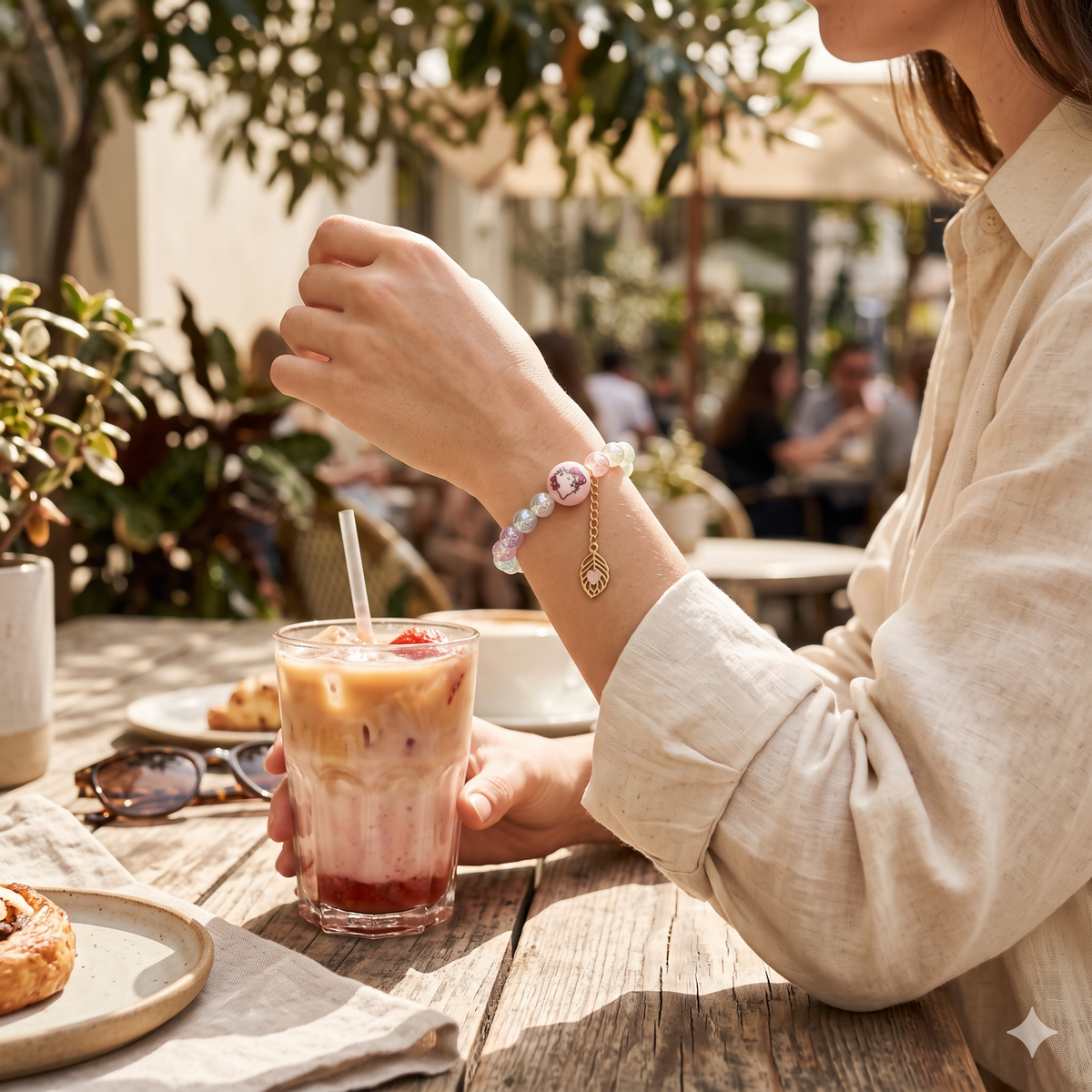 A person's arm wearing a pastel beaded bracelet with a pink Hello Kitty bead, holding an iced coffee at an outdoor cafe.