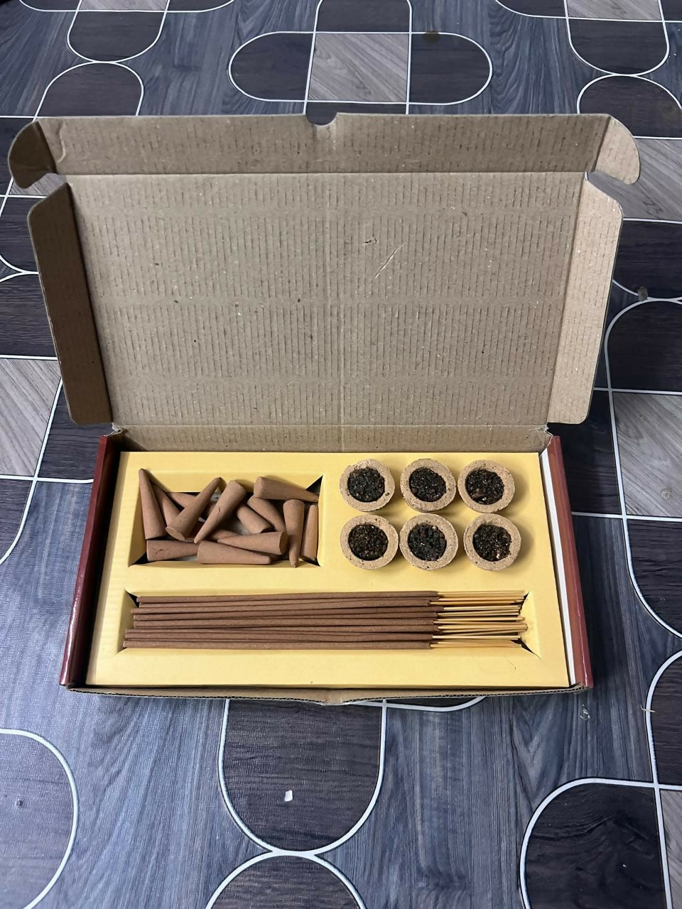An open cardboard box displaying dhoop cones, sambrani cups, and incense sticks on a dark patterned floor.