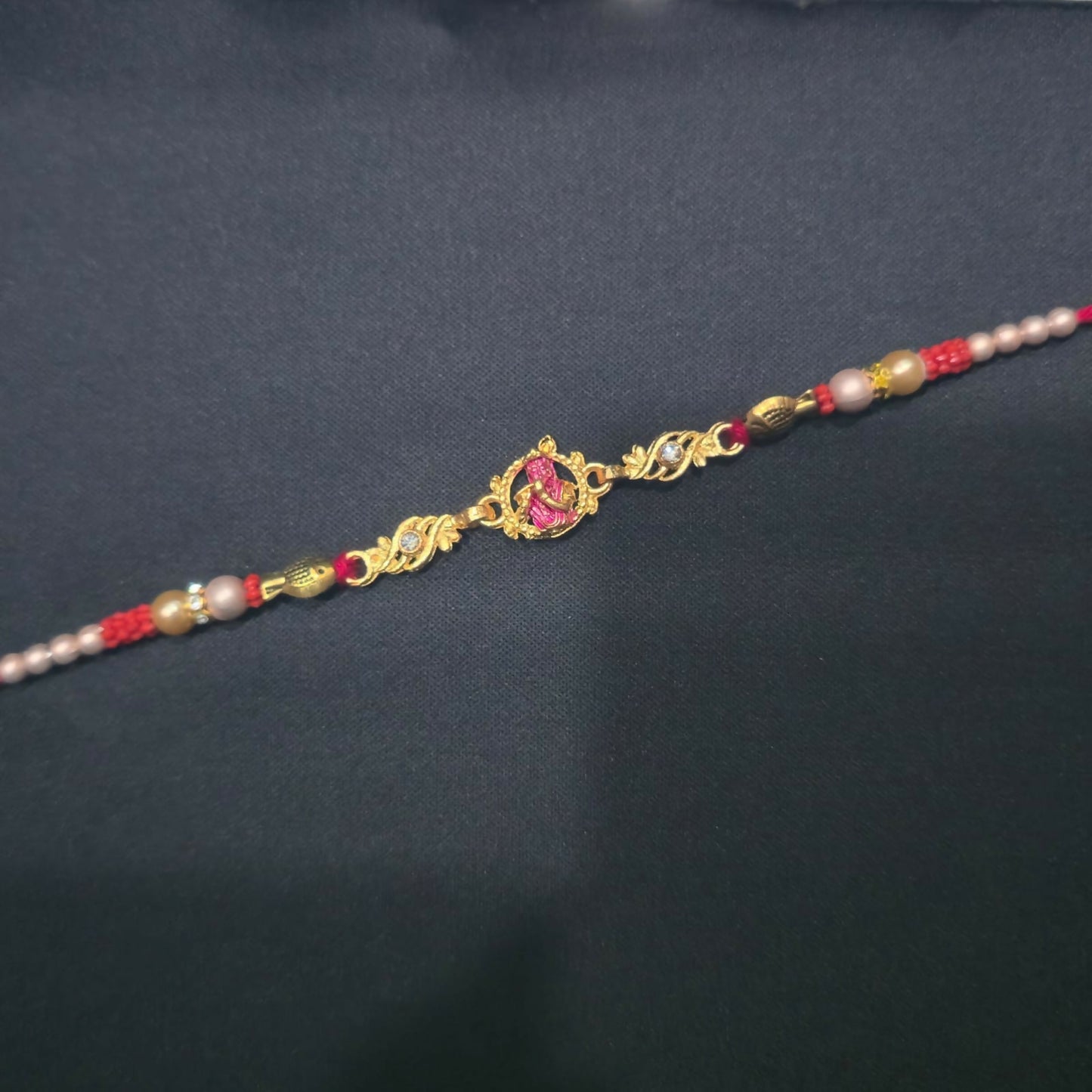 A gold and pink Lord Krishna Rakhi with red and white beads is centered on a plain black background.