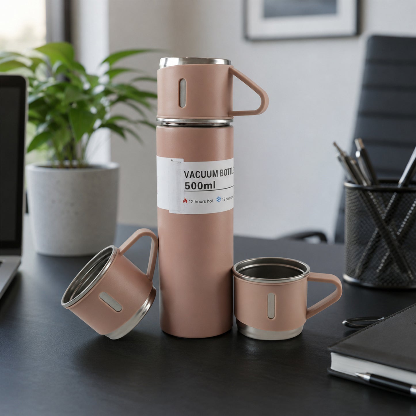 A pink vacuum flask and two matching cups are displayed on a dark office desk with a laptop, plant, and pen holder.