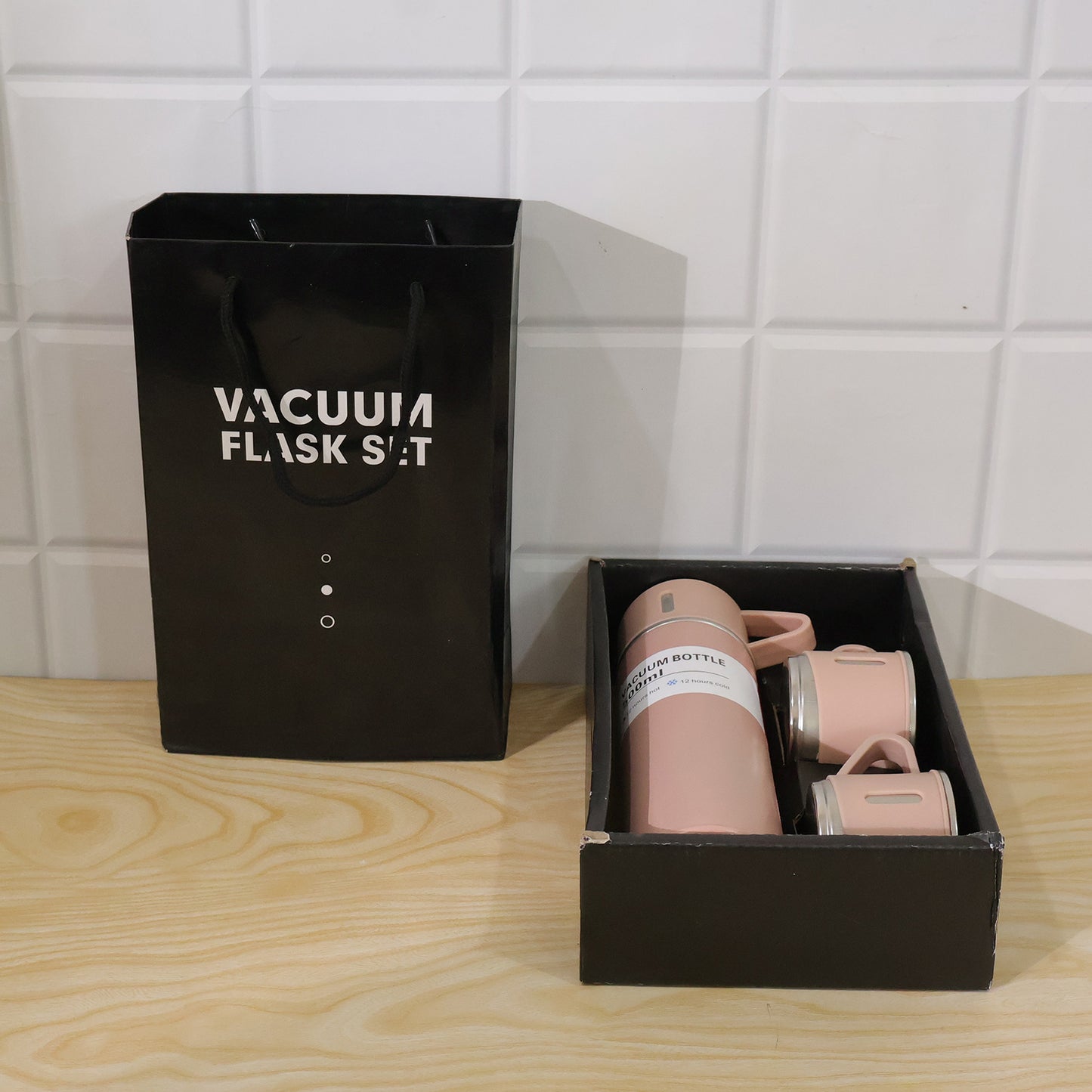 A black gift bag and a black box containing a pink vacuum flask set are placed on a wooden surface against a white tiled