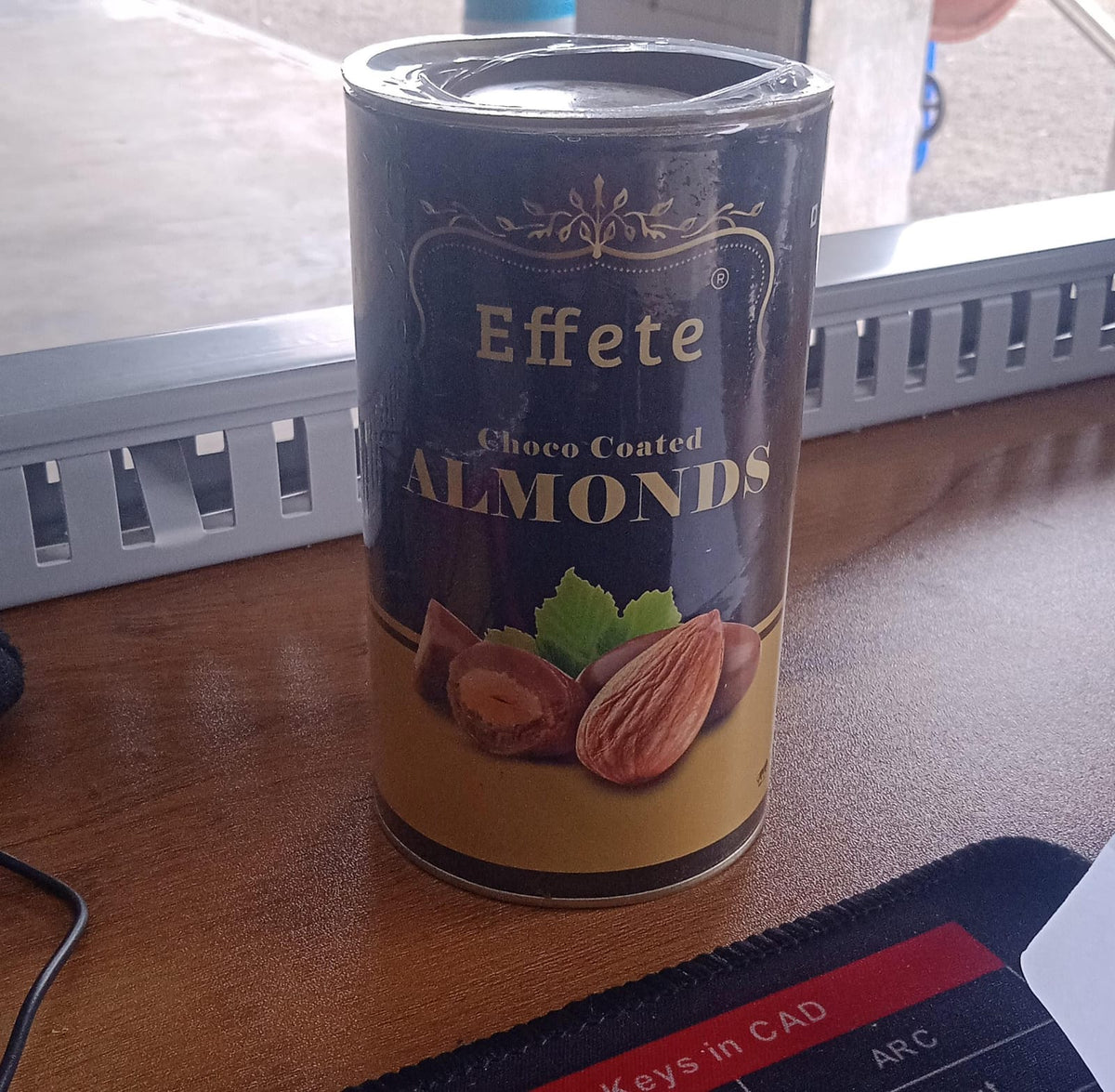 The Effete Choco Coated Almonds can is on a wooden desk with a black mouse pad and office items.