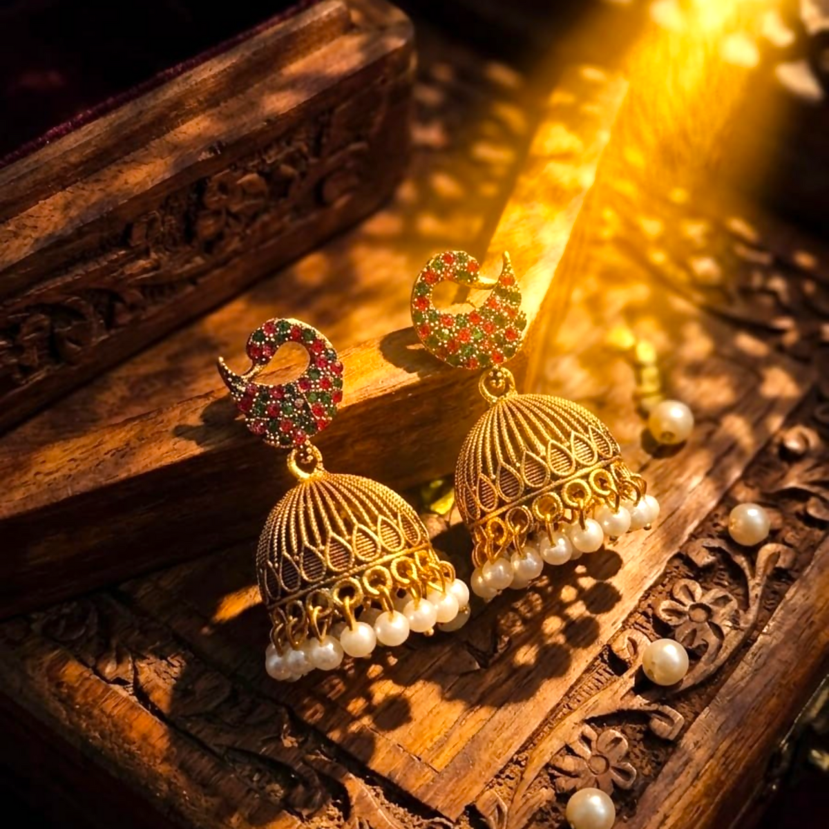 A pair of gold jhumka earrings with red and green stones and pearl drops on a wooden box.