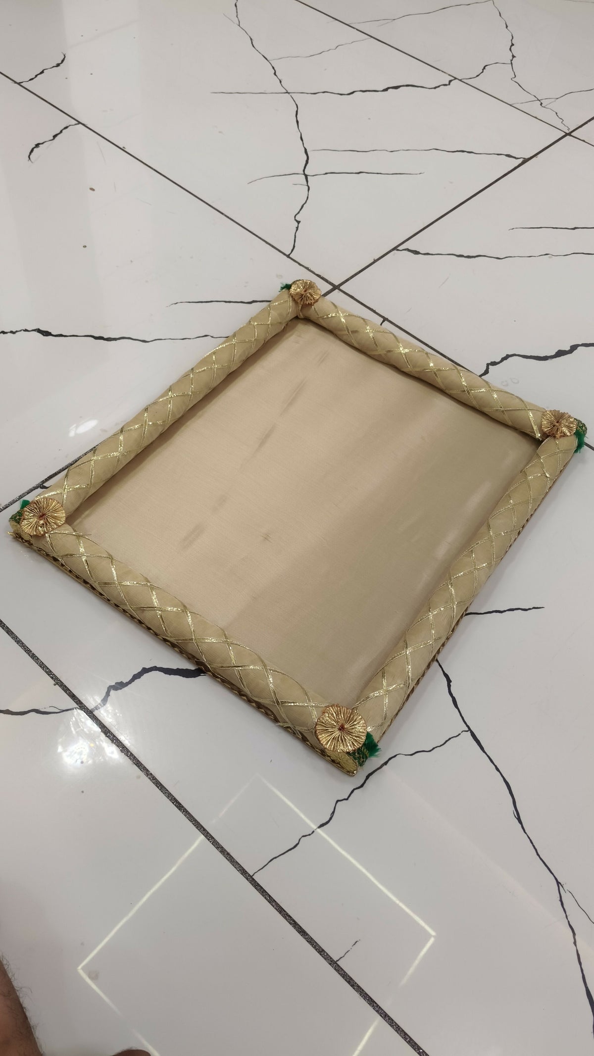 A simple beige fabric gift tray with gold braided edges and green corner accents, resting on a white tiled floor.