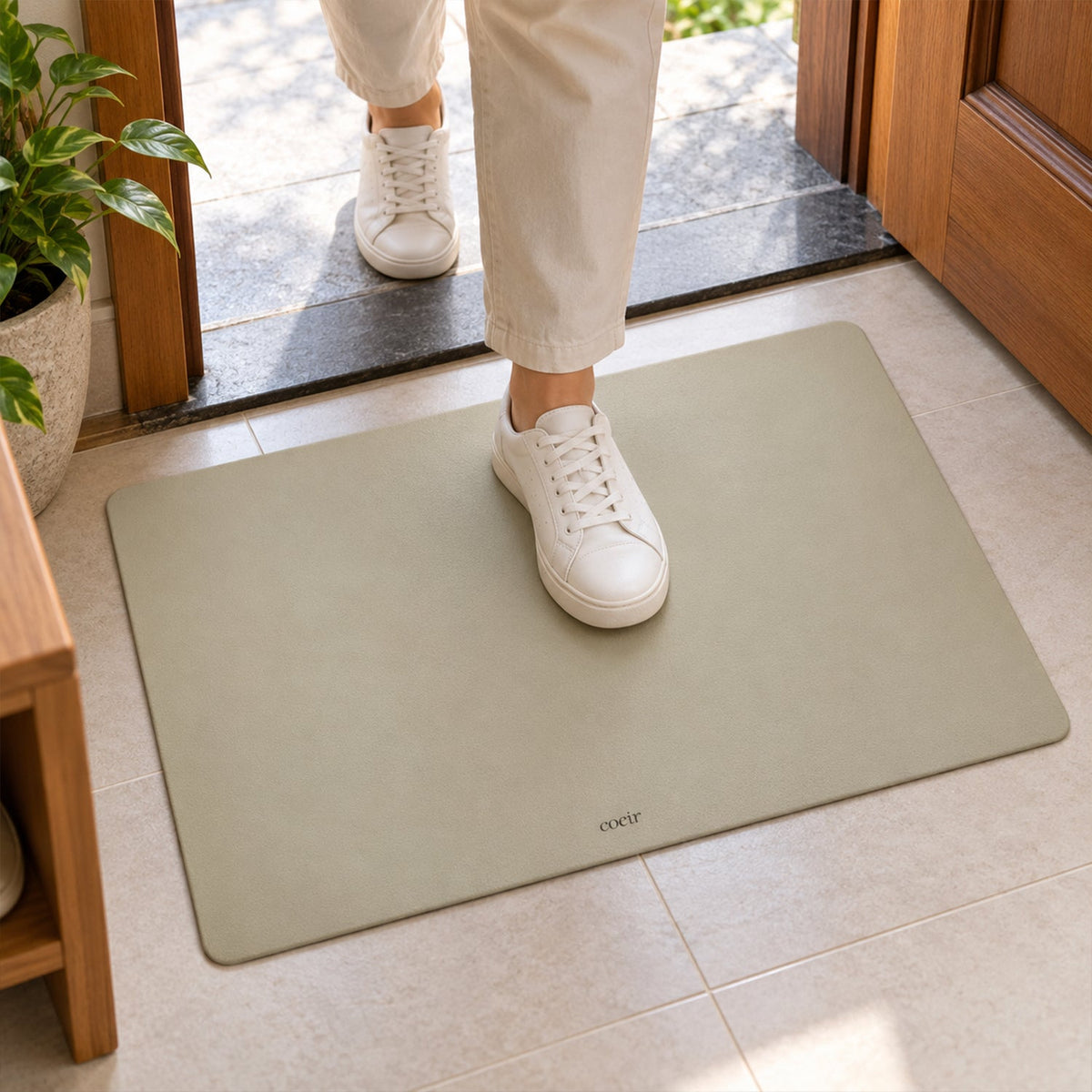 A person in white sneakers steps onto a light beige rectangular floor mat by a doorway.