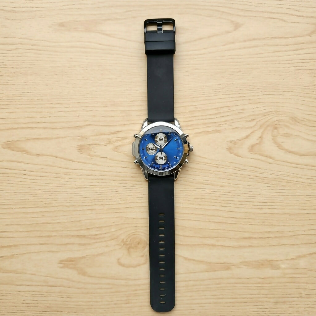 Top-down view of a black silicone strap watch with a silver case and blue dial, on a light wooden surface.