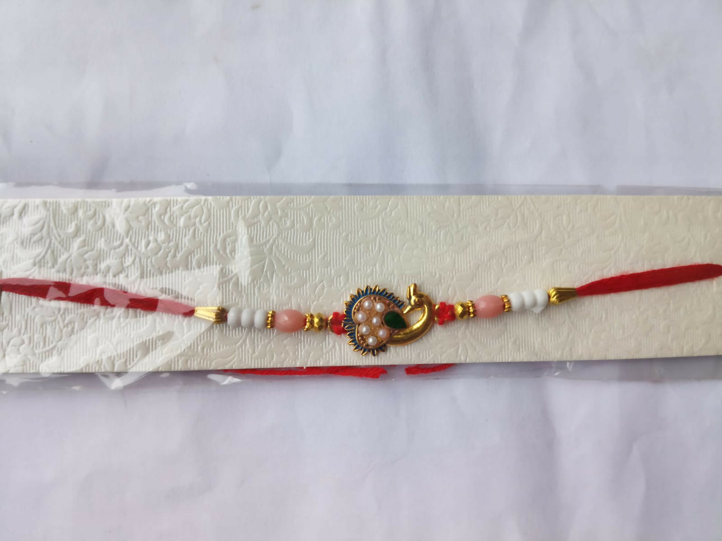 A red thread with a gold peacock charm, pink and white beads, and gold accents, displayed in clear packaging on a textured