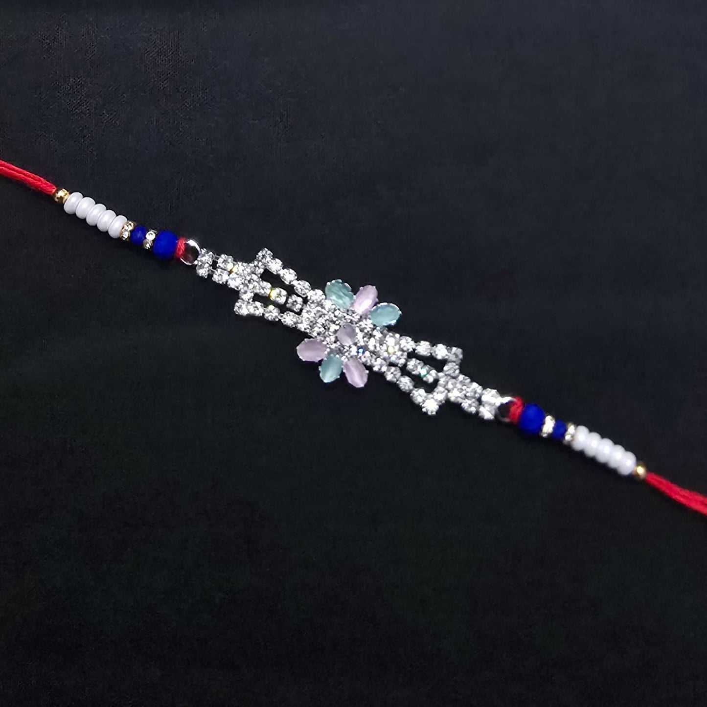 The elegant Rakhi, featuring a crystal floral design, blue beads, and red thread, is presented against a plain dark