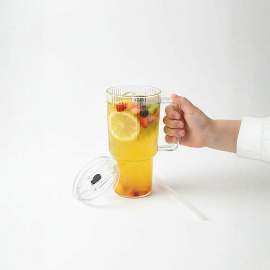 A hand holds a clear ribbed glass tumbler filled with a yellow drink and fruit slices, with the lid and straw beside it.