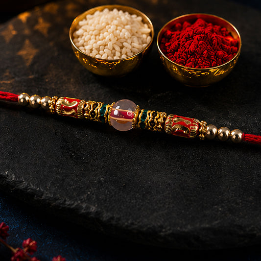 Close up of Golden Designer Rakhi featuring red and green stones with a gold-tone festive centre.