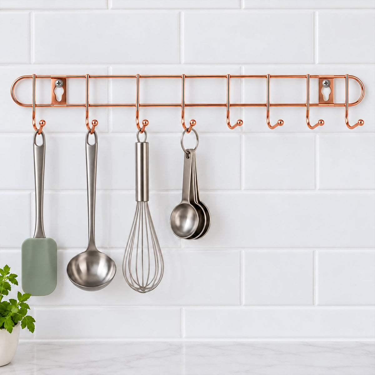 A rose gold metal wall hanger with eight hooks displays kitchen utensils against a white tiled background.