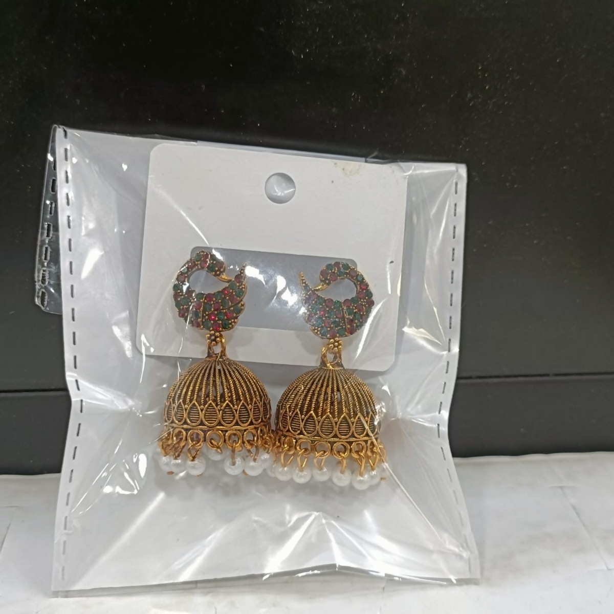 A pair of gold jhumka earrings with red and green stones and pearl drops in a clear plastic bag.