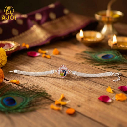 A silver mesh bracelet with a pink, green, and blue peacock charm lies on a wooden surface with peacock feathers and diyas.