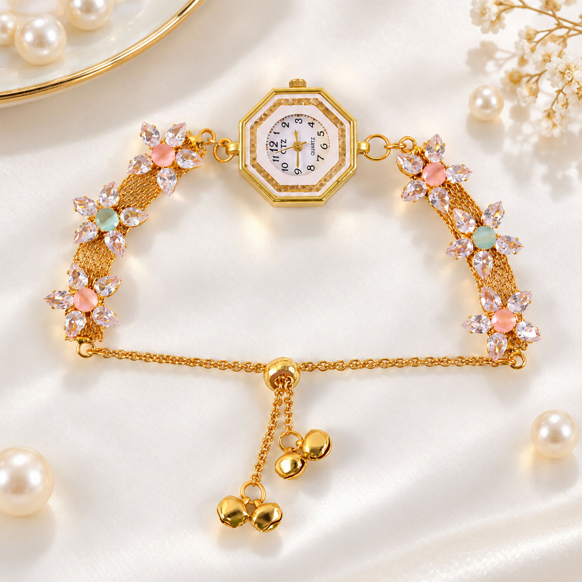 The gold-tone bracelet watch with crystal flowers and dangling bells is displayed on a white satin surface with pearls.