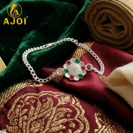 Silver-plated Krushna Rakhi bracelet with a flower-shaped centerpiece adorned with red and green stones, resting on red and
