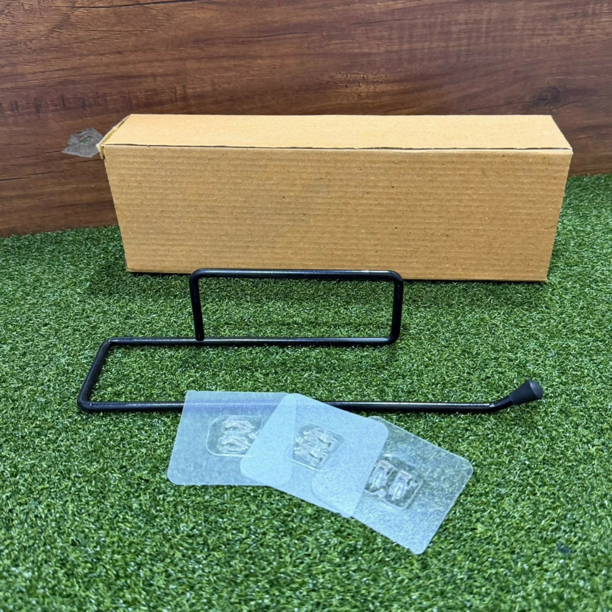 Disassembled black metal paper towel holder with clear adhesive pads and a brown cardboard box on green grass.