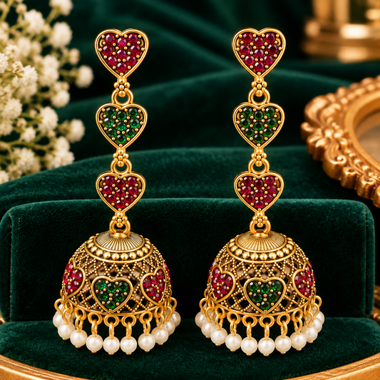 A pair of ornate gold-toned jhumka earrings with red and green heart-shaped stones and pearl drops, resting on green velvet.