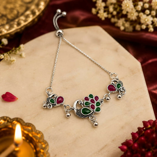 Silver bolo bracelet with ruby and emerald peacock motifs, surrounded by traditional decor.