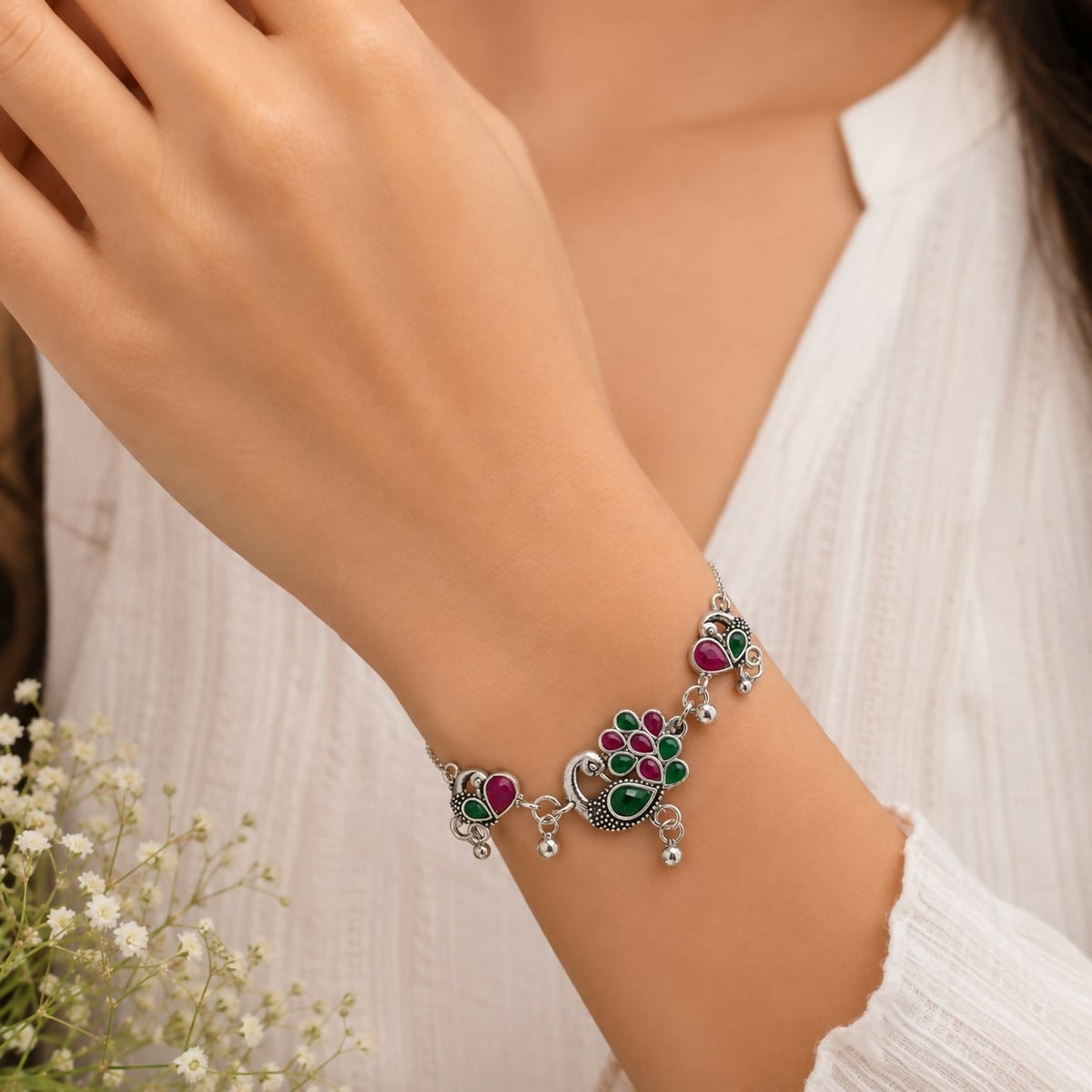A woman's hand wearing a silver bolo bracelet with ruby and emerald peacock motifs.