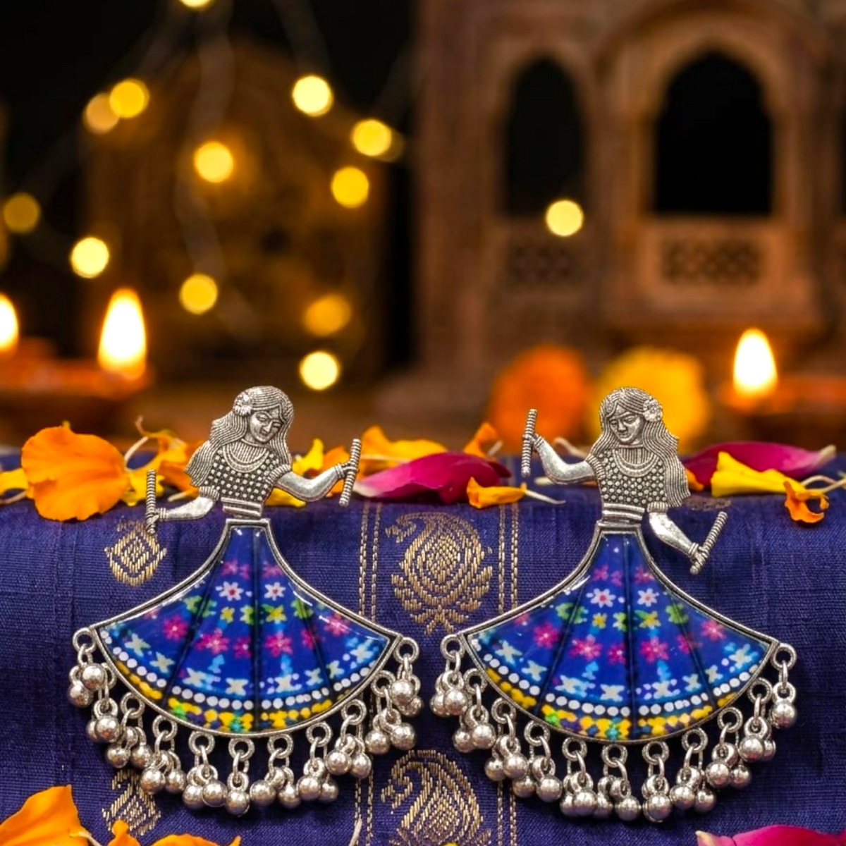Pair of oxidized silver dancing doll earrings with blue floral skirts on a purple textile with warm lights.