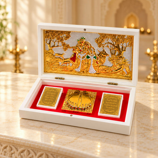 An open white wooden box reveals a gold-toned Radha Krishna scene, two mantra plaques, and a jeweled Charan Paduka on red