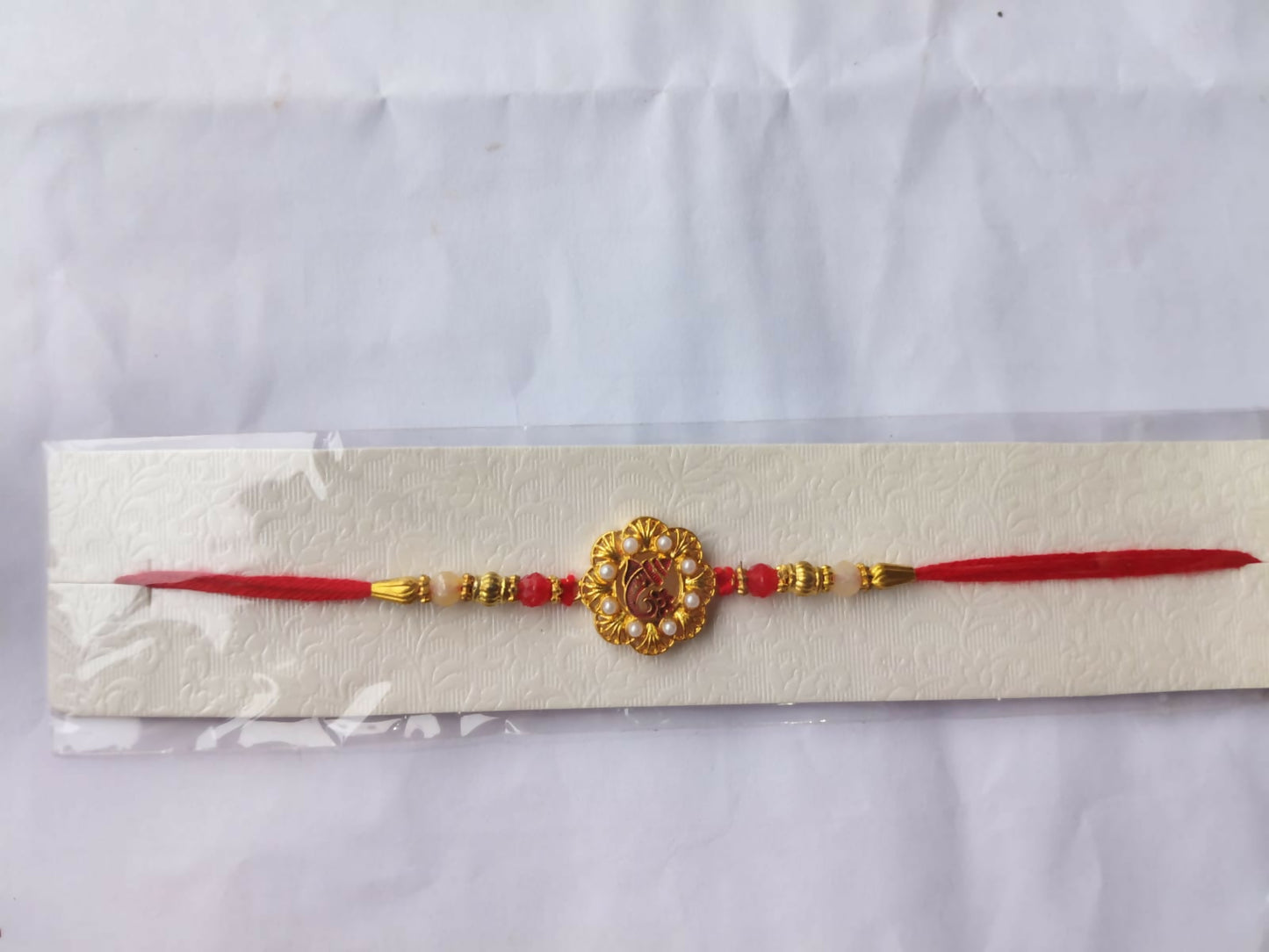 A red and gold Ganesha bracelet with red beads and golden detailing is displayed on a textured white card inside a clear
