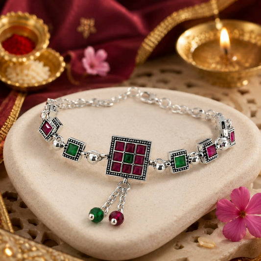 Antique silver bracelet with ruby-red and emerald-green square inlays on a stone surface with a lit candle.