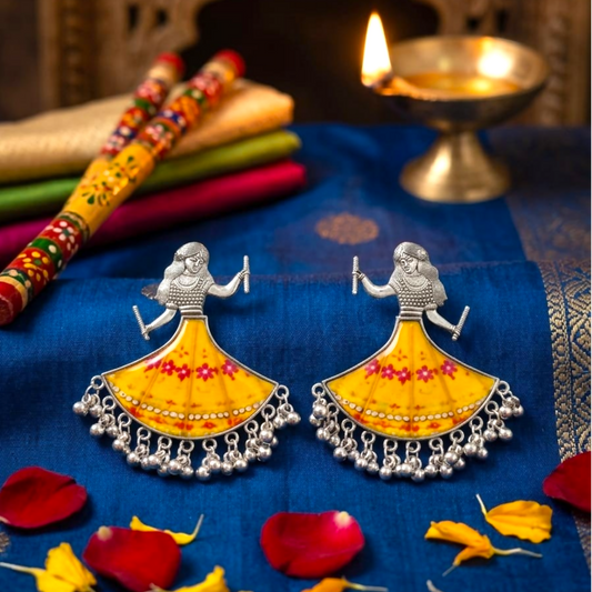 A pair of silver-toned earrings with a yellow skirt design and small bells on a blue fabric with a lamp.
