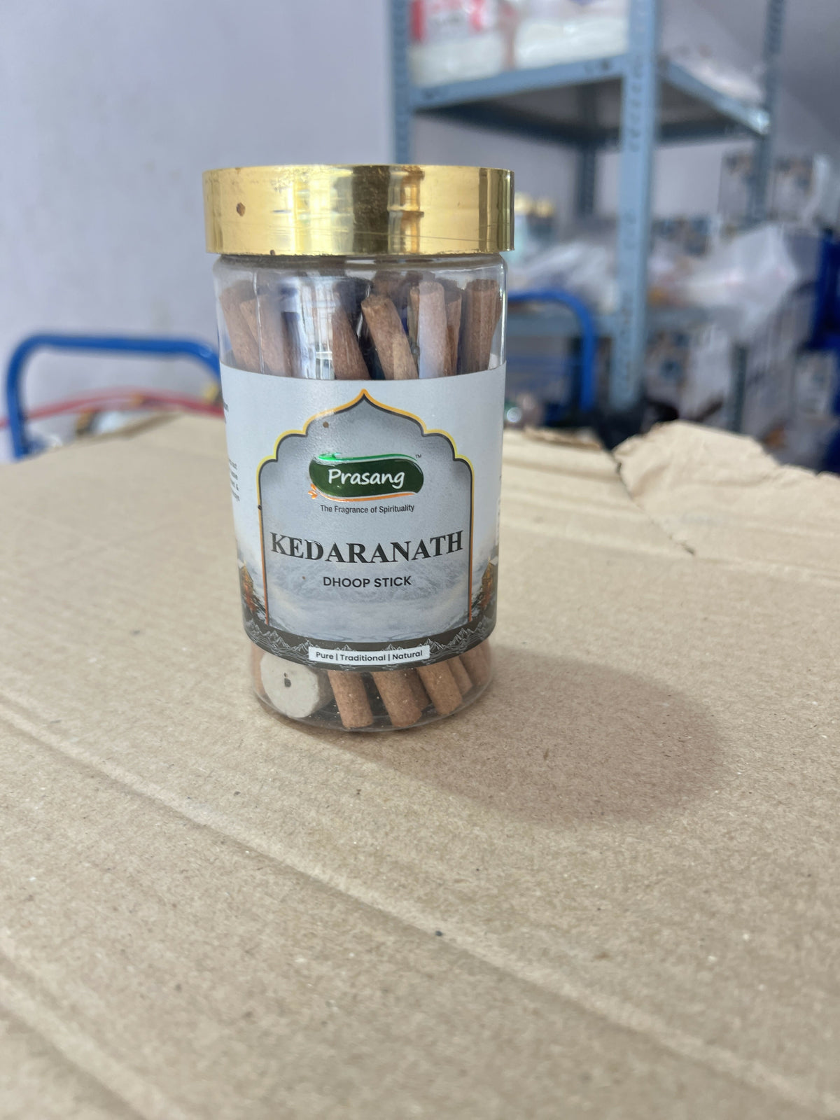 A clear plastic jar with a gold lid, filled with brown dhoop sticks, sitting on a cardboard box.