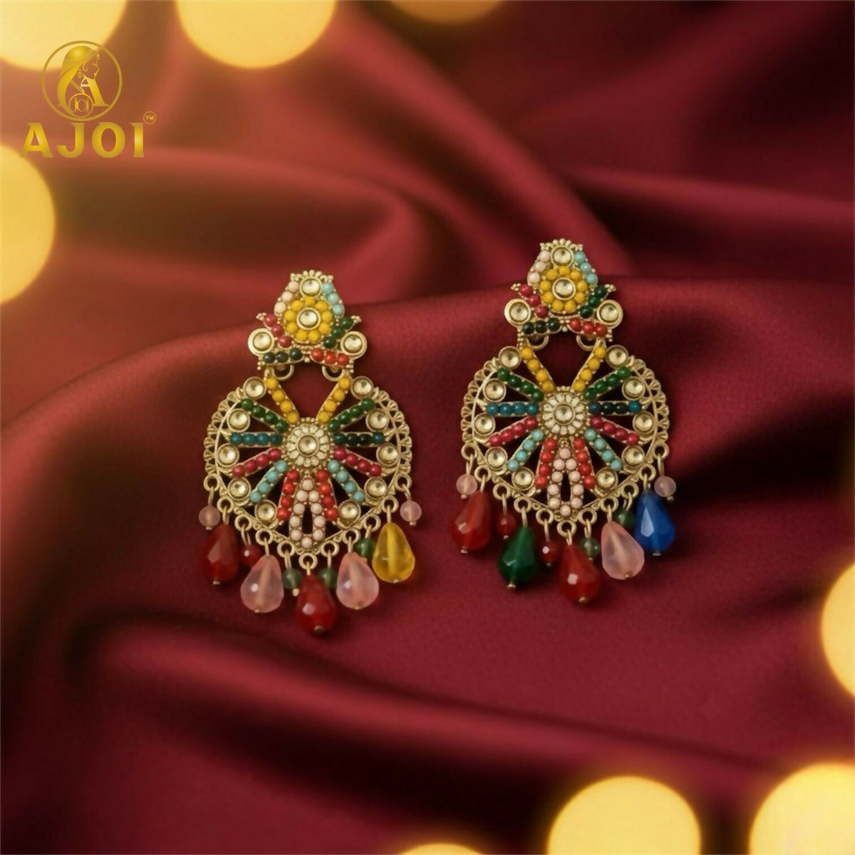 A pair of festive gold earrings with bright beads and colorful teardrop accents rests on a rich red fabric.