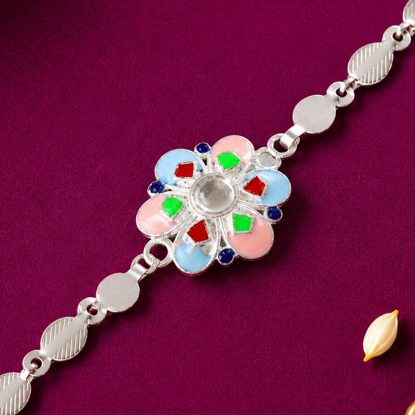 A silver chain rakhi with a colorful enamel flower charm on a deep purple background with a rice grain.