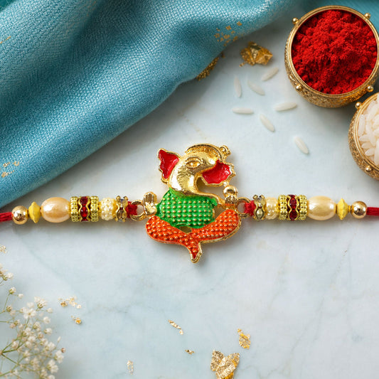 A Ganesha charm with red, green, and gold enamel sits on a red thread with pearl and gold beads, on a light blue fabric with