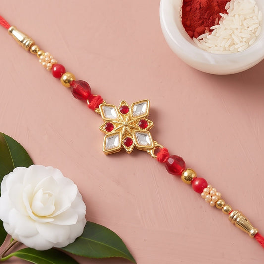 A gold flower-shaped charm with clear and red gems on a red thread with red and gold beads, next to a white camellia and a
