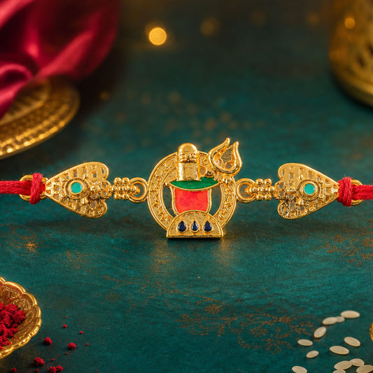 A gold-toned Shivling Trishul charm with red, green, and blue enamel work sits on a red thread against a dark teal