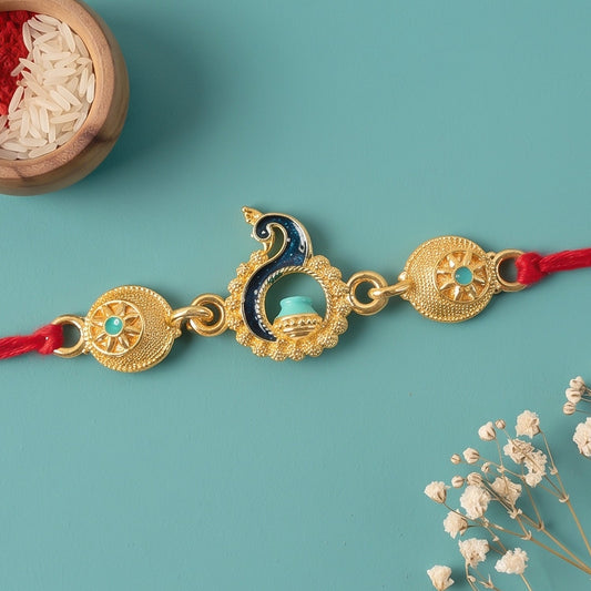 A gold-toned peacock Rakhi with turquoise enamel and red cord lies on a teal surface next to a small wooden bowl of rice.