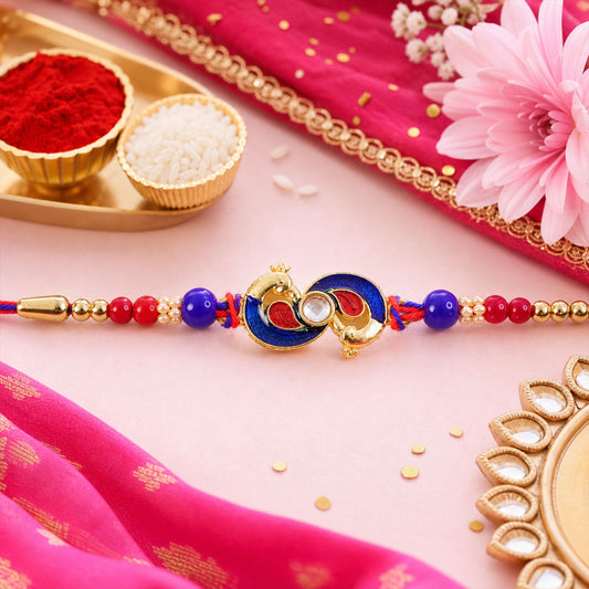 A gold-colored peacock-shaped charm with blue and red enamel, flanked by red and blue beads, lies on a pink fabric with