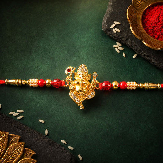 A gold-colored Ganpati charm with clear crystals and red beads is centered on a red thread, resting on a dark green surface